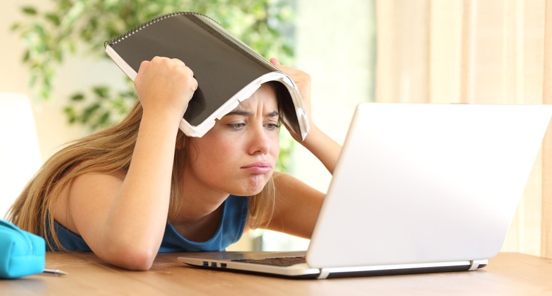 A frustrated young woman holds a notebook over her head while staring at a laptop screen.