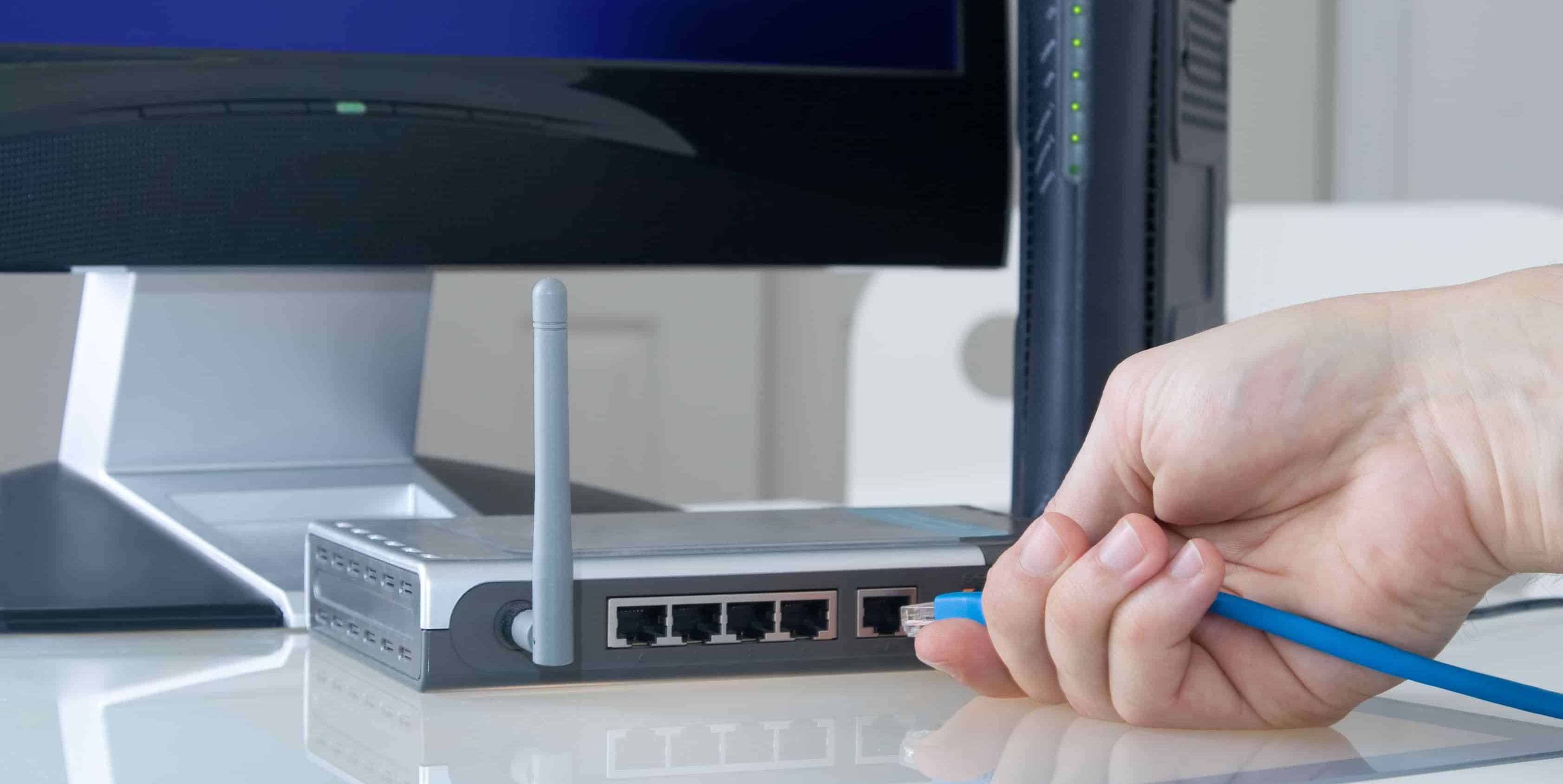  A hand plugging a blue Ethernet cable into a Wi-Fi router, with a computer monitor in the background.