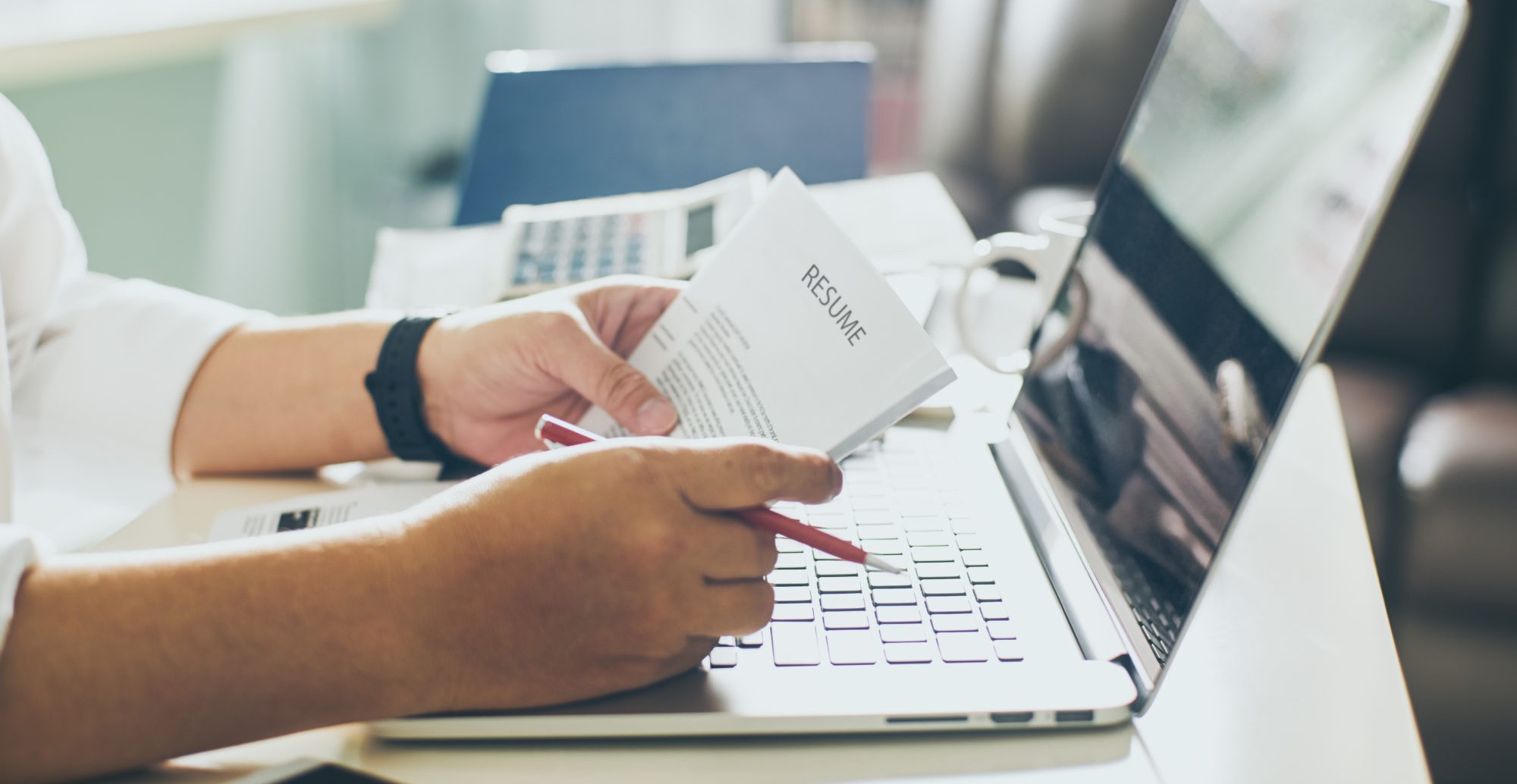 A person holding a resume and a pen while reviewing it in front of a laptop on a desk.