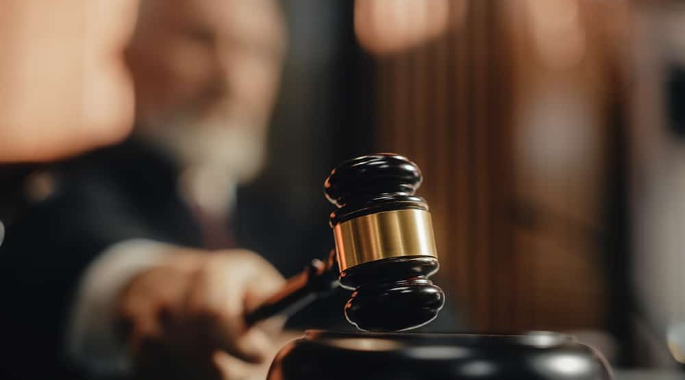  A judge's hand holding a wooden gavel, poised to strike, with a blurred courtroom background.